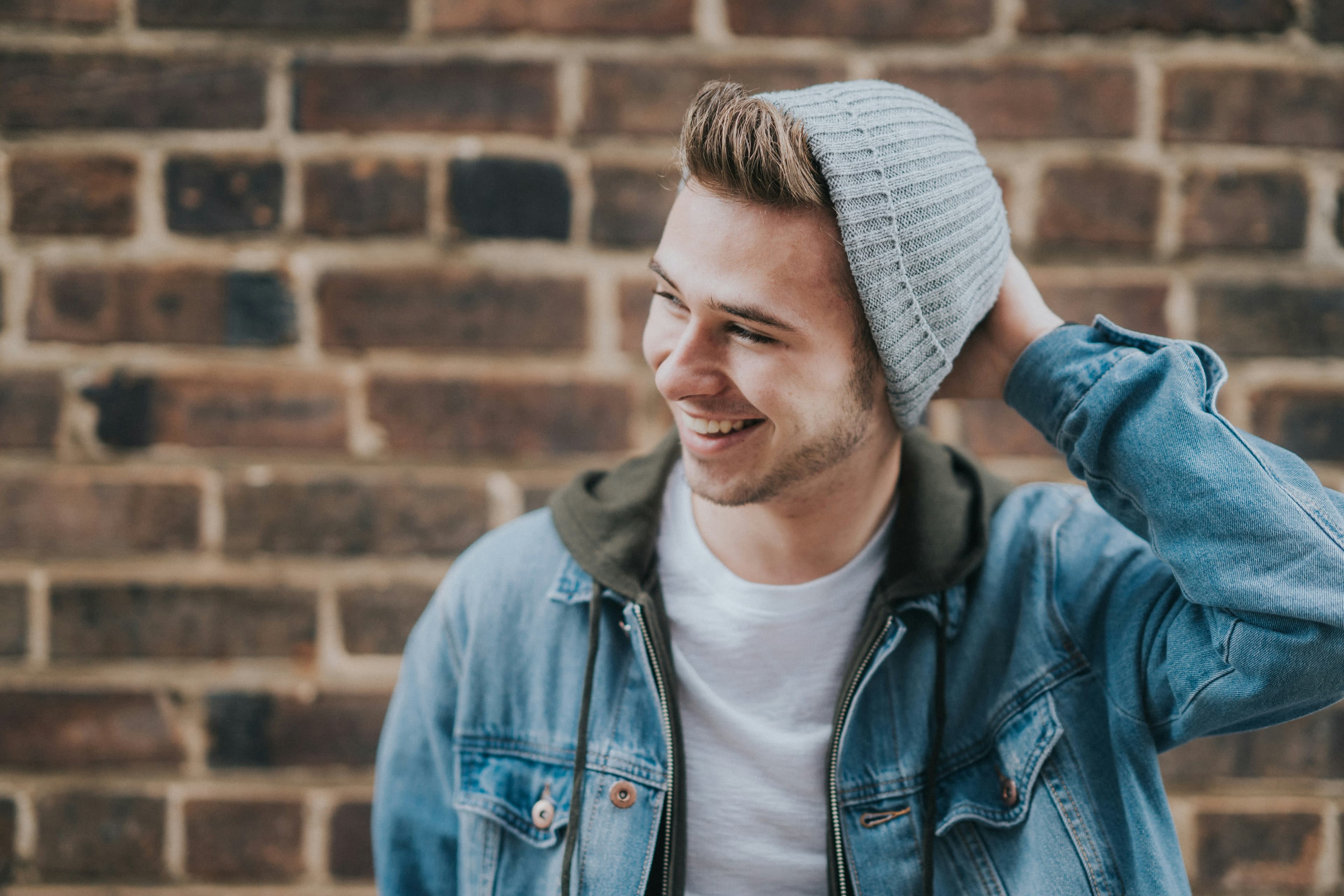 A smiling young man in a denim jacket and beanie leans against a brick wall, adjusting his hat.