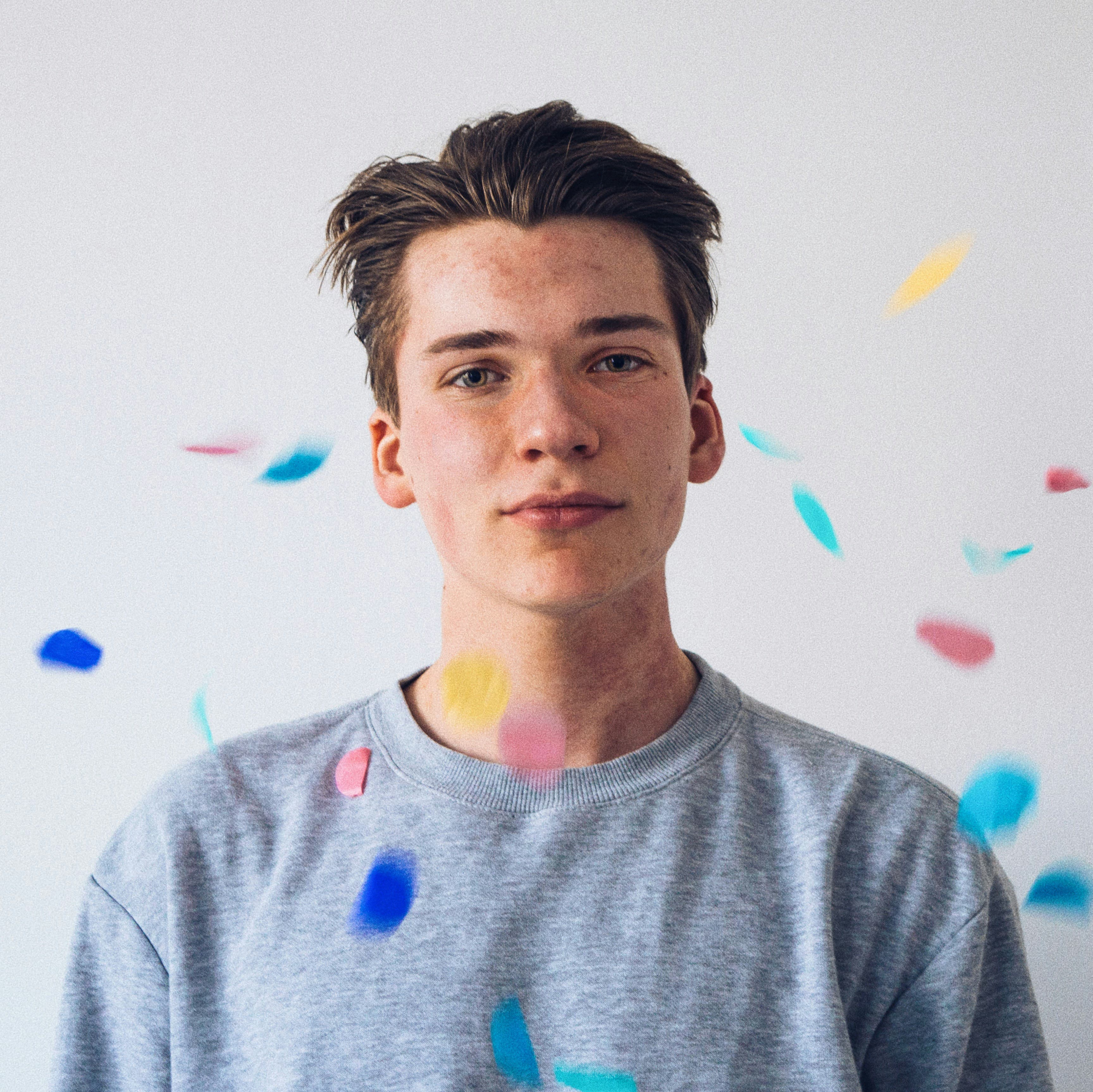 A young man in a gray sweatshirt stands against a white background with colorful confetti floating around him.