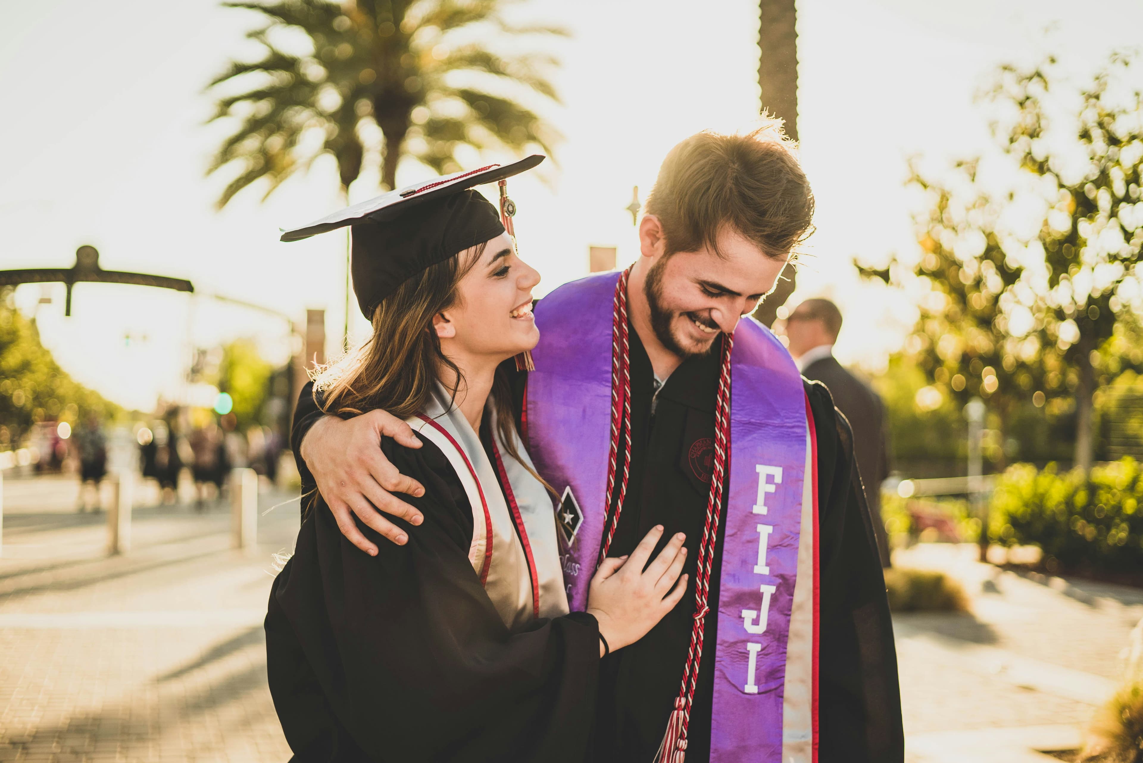 Two graduates in caps and gowns embrace and smile in the sunlight, celebrating their achievement.