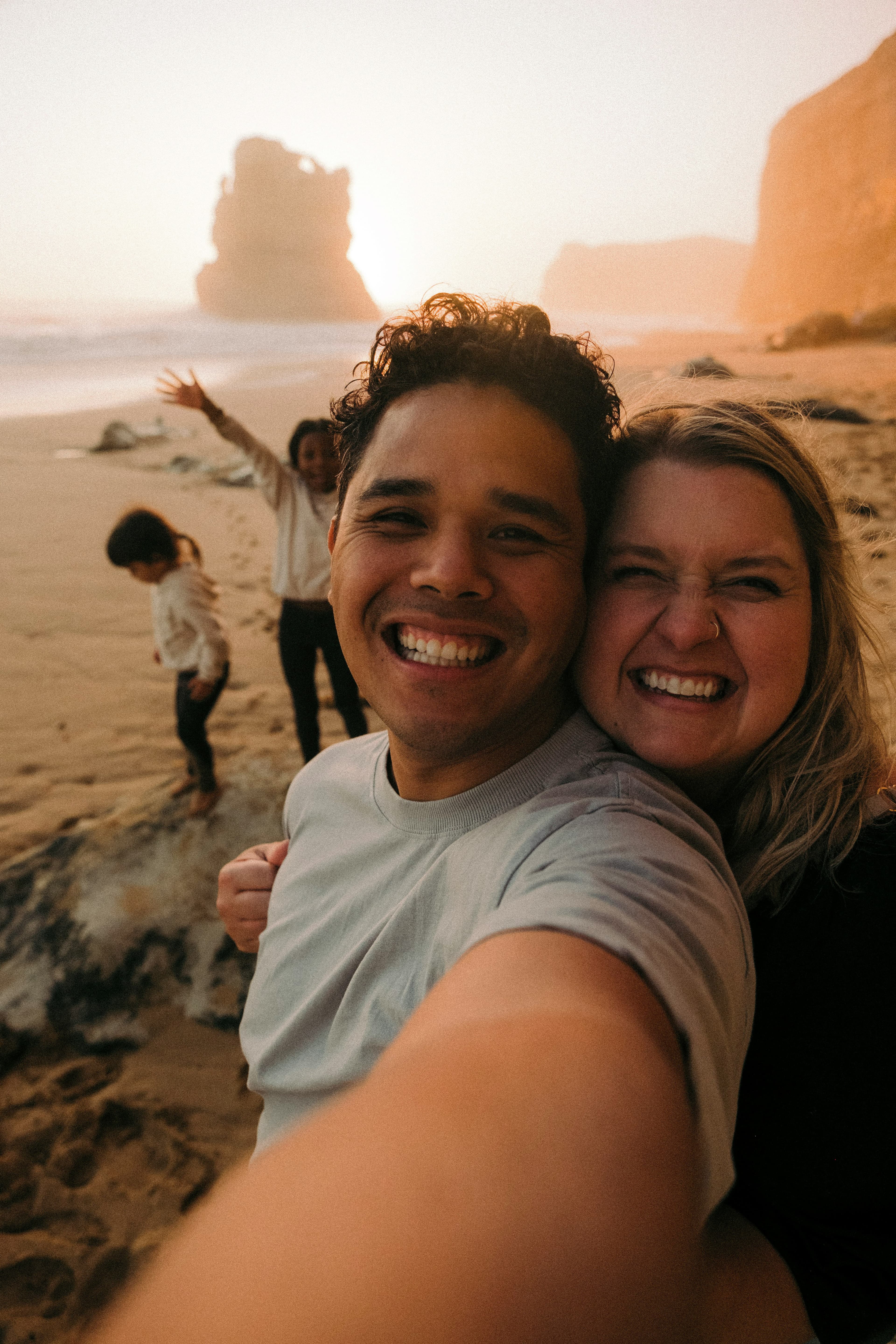 A smiling couple takes a selfie on a beach at sunset, with two children playing in the background.