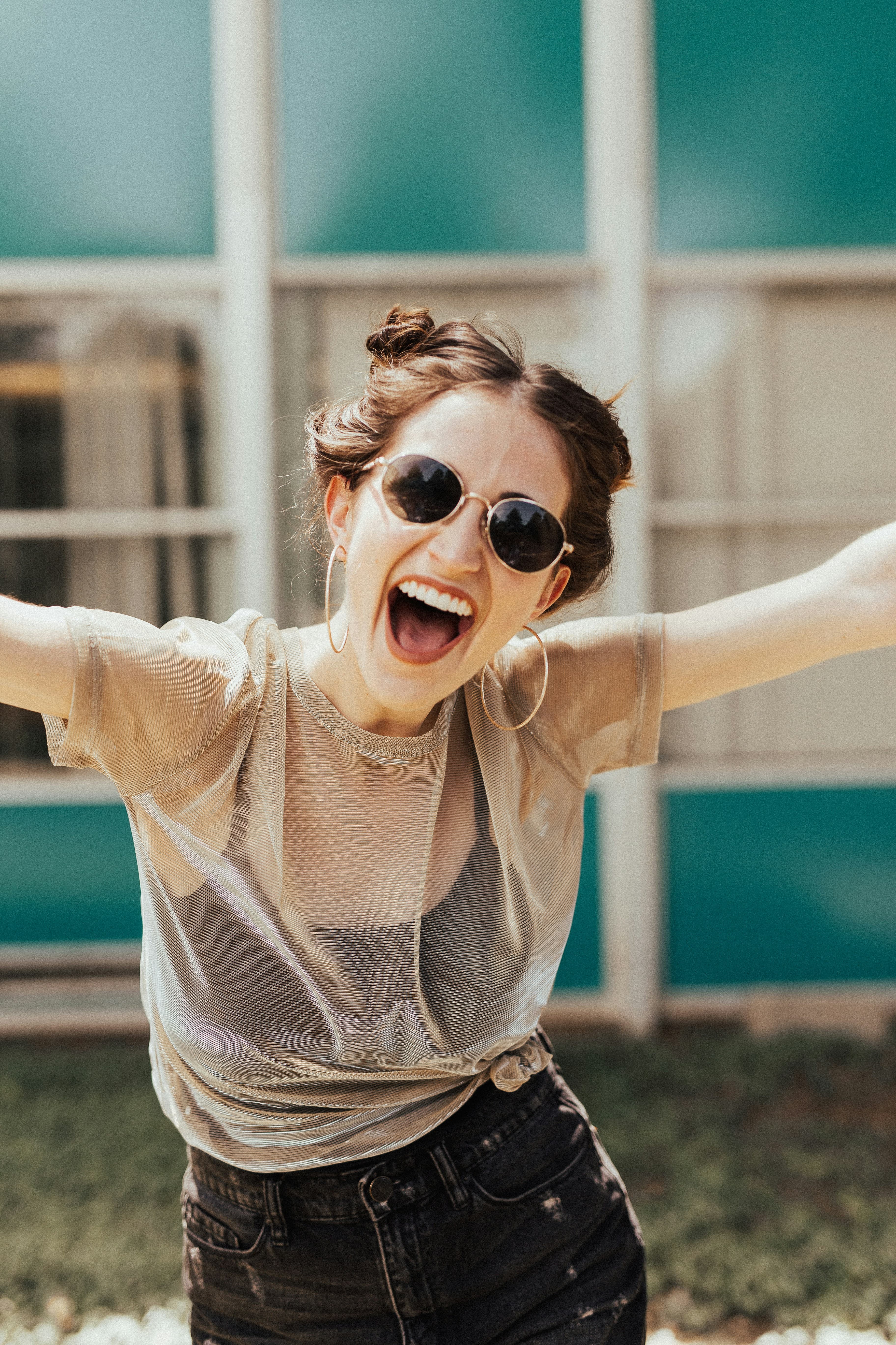 A joyful young woman with sunglasses, hoop earrings, and a sheer top, smiling with arms outstretched outdoors.